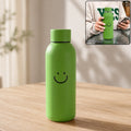 Green glass water bottle with a smiley face on a wooden table, with an inset showing a person holding a similar bottle.