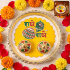 A gold-rimmed white and yellow pooja thali with Radhe Radhe text and two small containers is surrounded by orange and yellow
