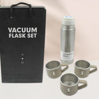 A black gift bag and a silver vacuum flask with three beige mugs are displayed against a softly blurred background.