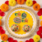 A silver and yellow Radhe Radhe pooja thali set with two small decorative containers, surrounded by marigolds and rose