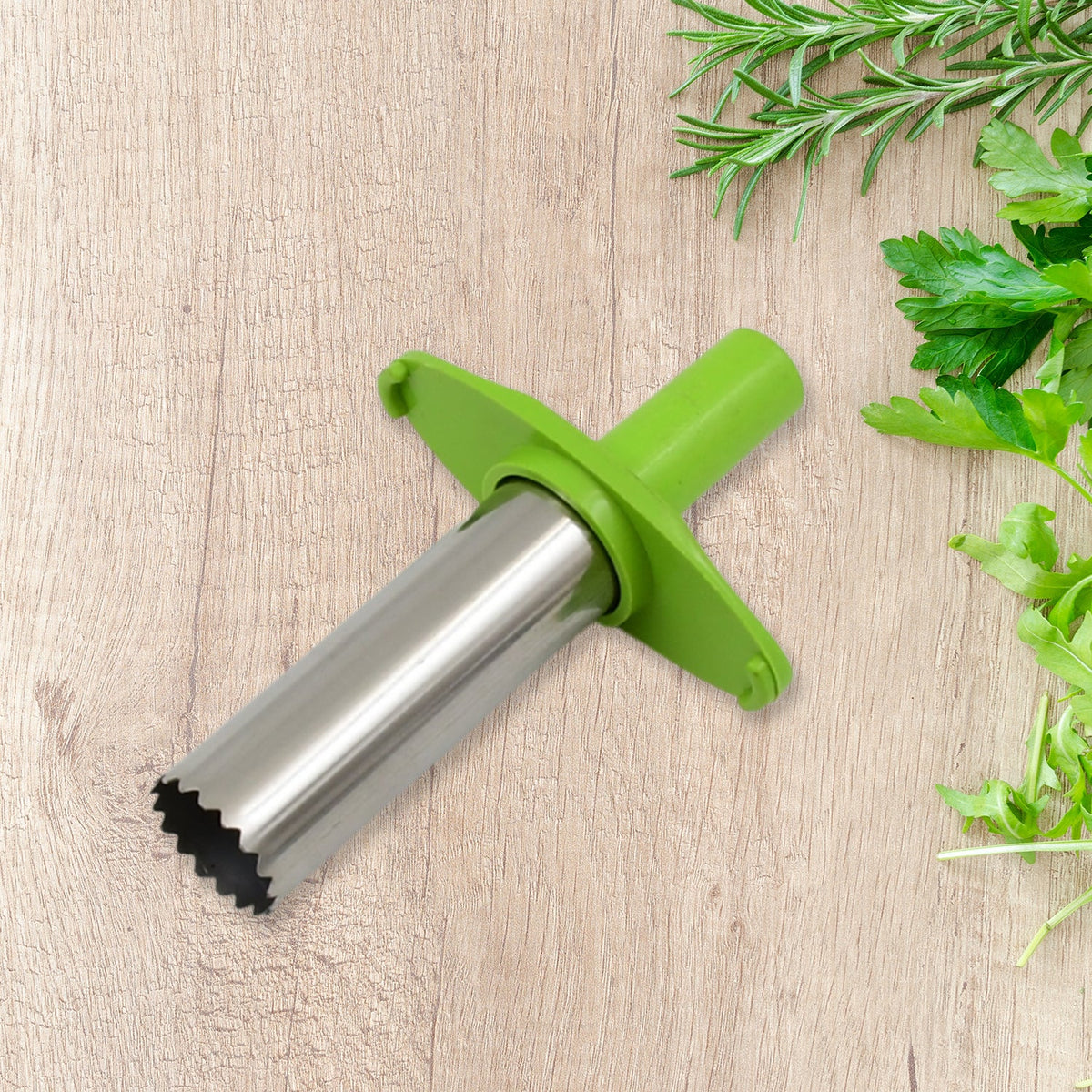 A green and silver chili corer rests on a wooden surface, surrounded by fresh green herbs like rosemary and parsley.