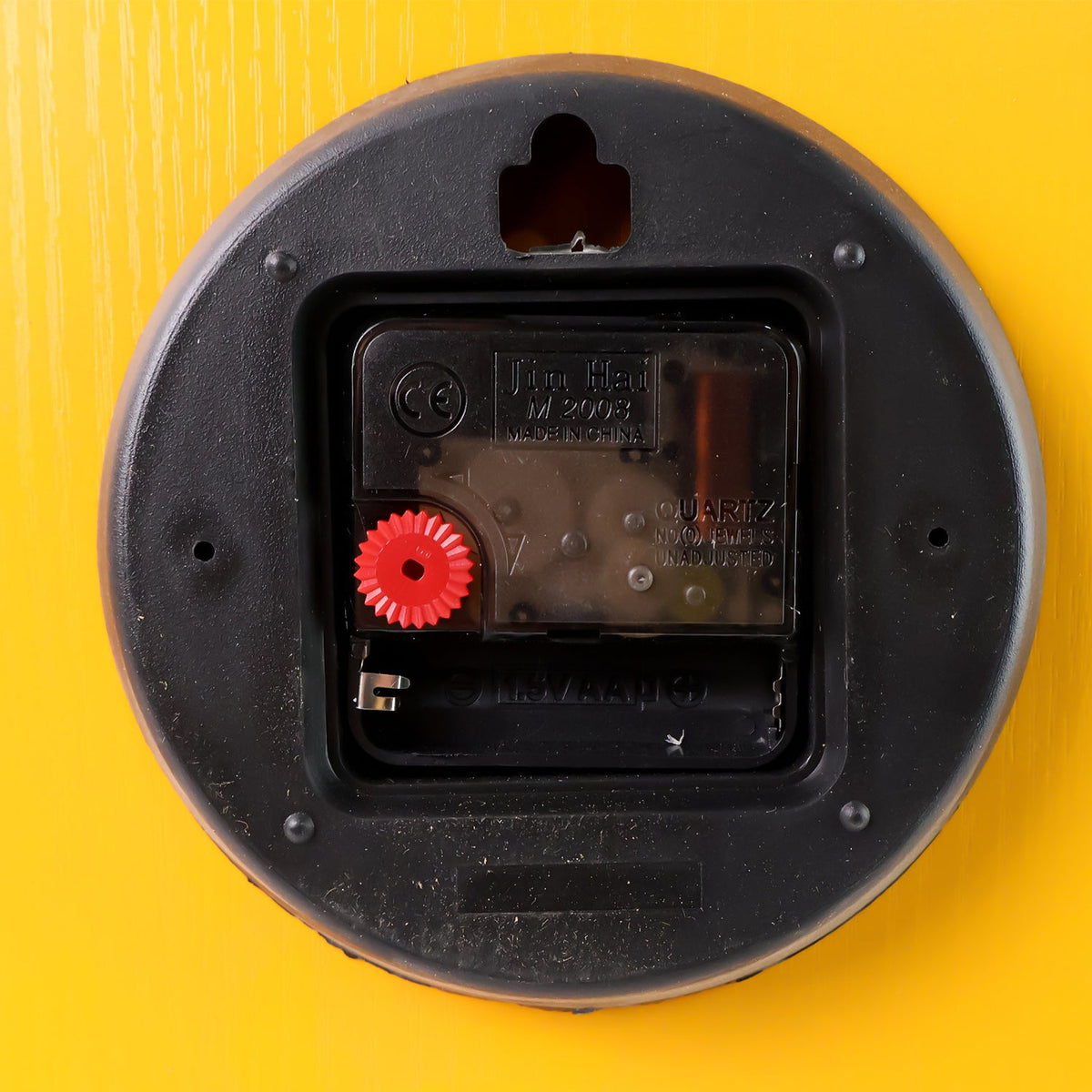 The black plastic back of a quartz clock mechanism with a red adjustment knob is visible against a yellow background.
