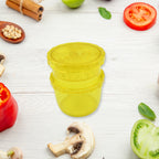 A yellow plastic manual food grinder is centered on a white wooden surface, surrounded by various fresh vegetables and