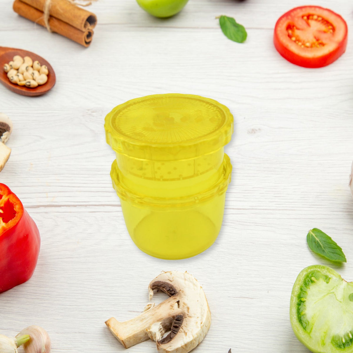 A yellow plastic manual food grinder is centered on a white wooden surface, surrounded by various fresh vegetables and