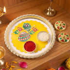 A gold-rimmed white and yellow pooja thali with Radhe Radhe text holds small containers of rice and red powder, with two