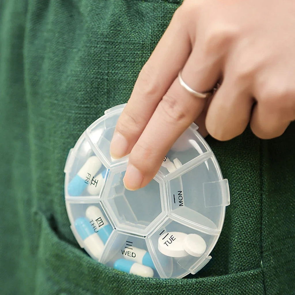 A hand placing a clear 7-day pill organizer with blue and white pills into a green pocket.