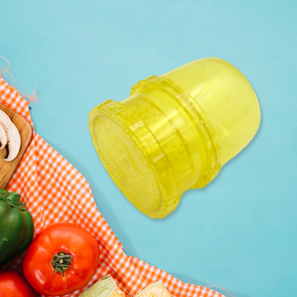 A yellow plastic manual food grinder is angled on a blue background, with a red and white checkered cloth and vegetables in