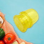 A yellow plastic manual food grinder is angled on a blue background, with a red and white checkered cloth and vegetables in