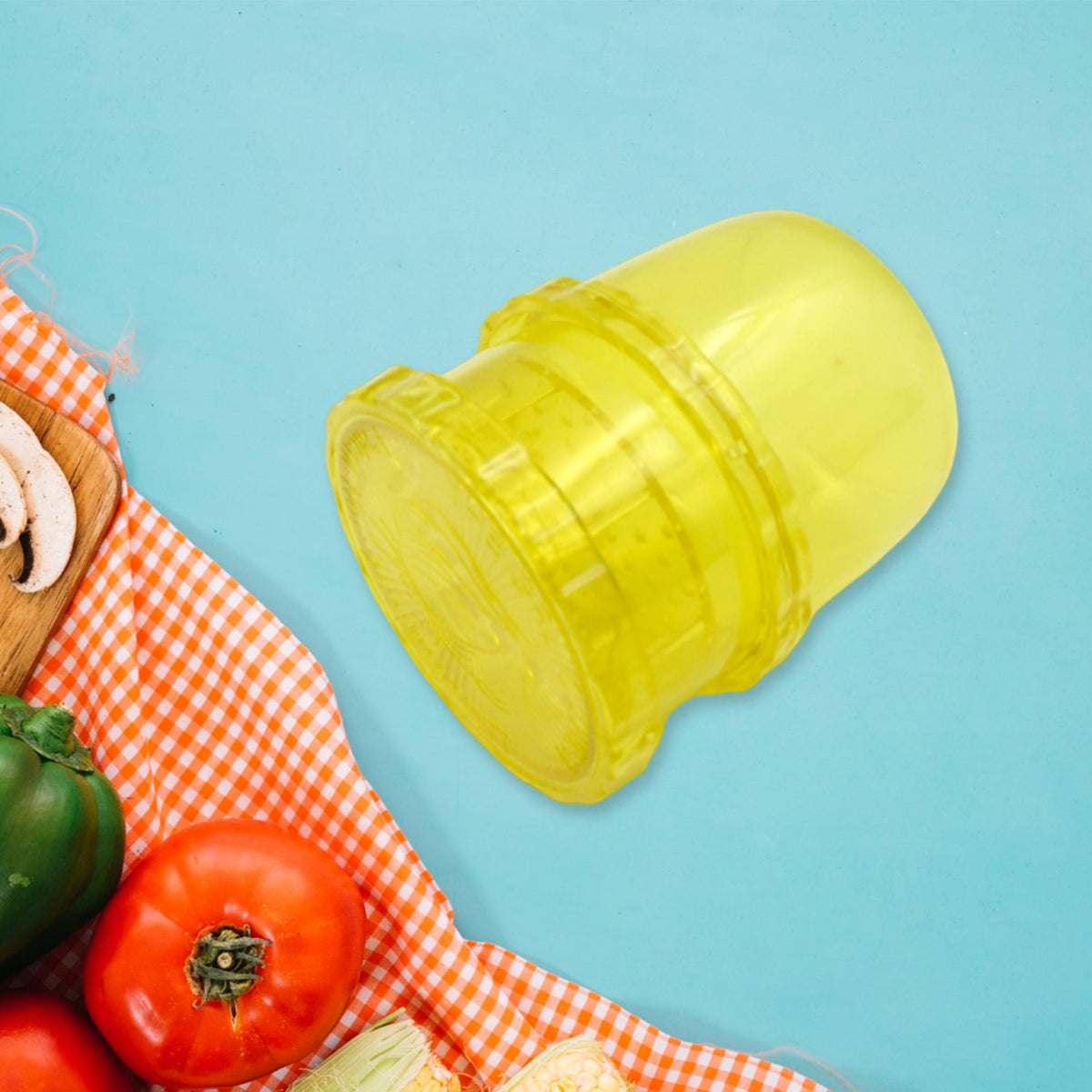 A yellow plastic manual food grinder is angled on a blue background, with a red and white checkered cloth and vegetables in