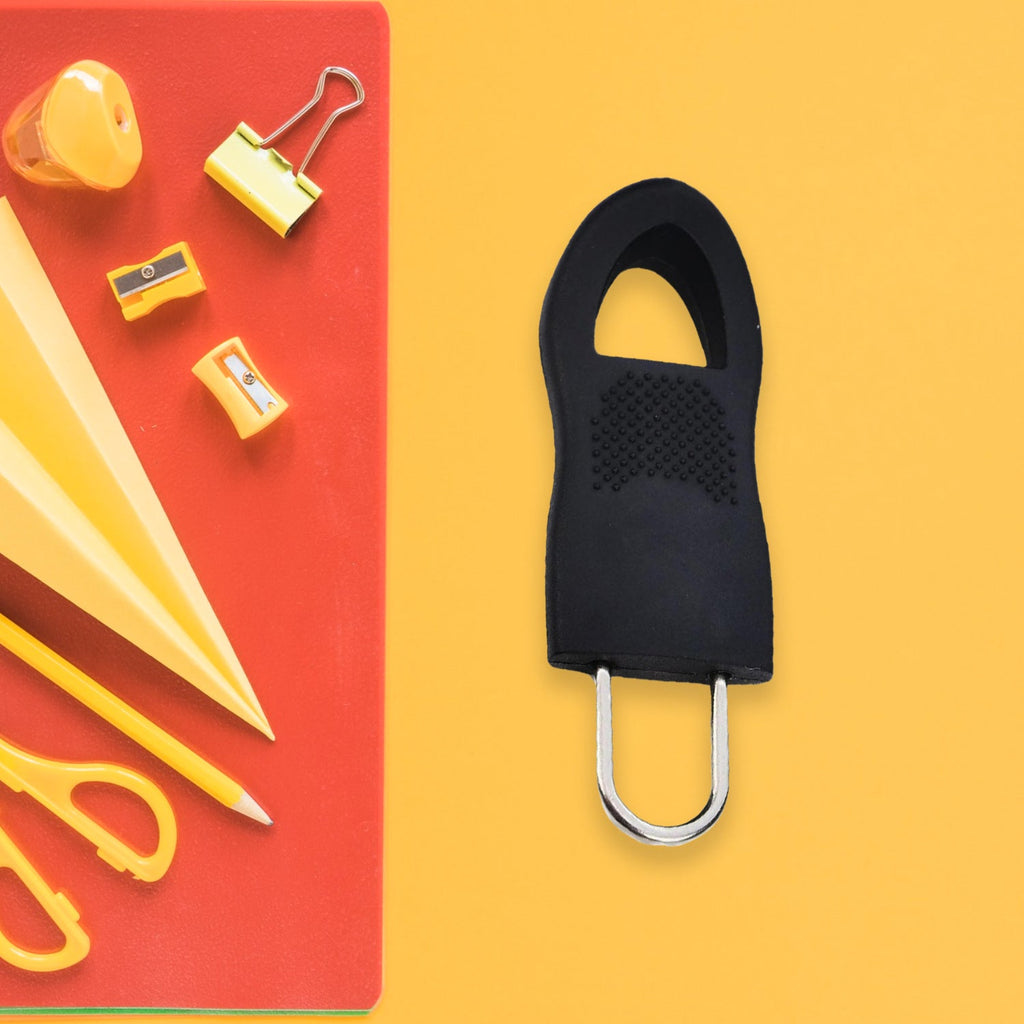 A black rubber zipper pull tab with a silver metal loop is on a yellow surface next to a red notebook.