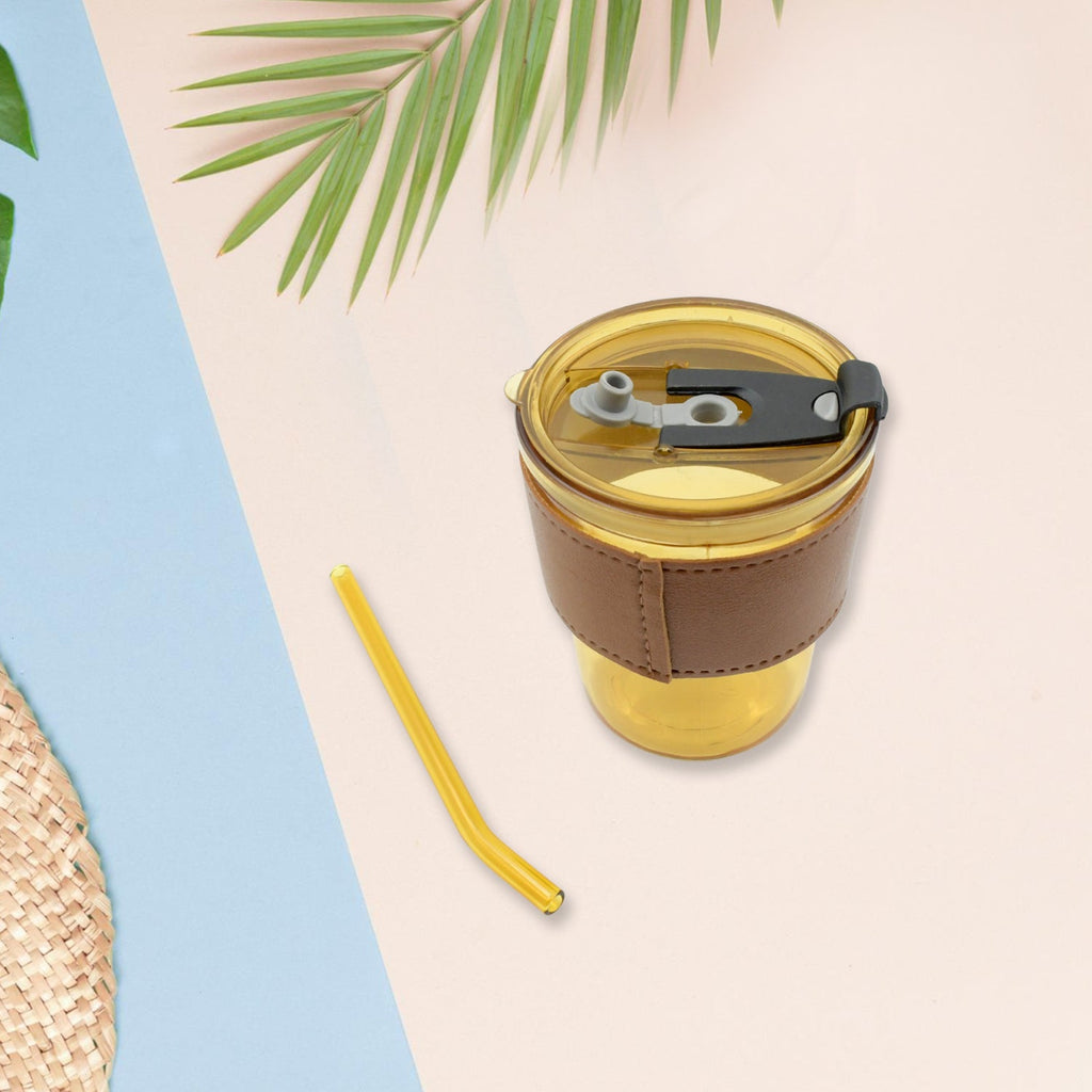 An amber glass cup with a brown leather sleeve and a separate straw are on a pink and blue background with palm leaves.