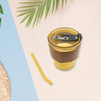 An amber glass cup with a brown leather sleeve and a separate straw are on a pink and blue background with palm leaves.