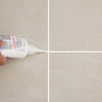 Close-up of a hand applying white sealant from a tube to the intersection of four beige floor tiles.