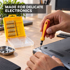 A person uses a yellow and red screwdriver from a 32-piece set to repair delicate electronics on a wooden desk.