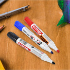 Three whiteboard markers in black, blue, and red, with their caps on, lying on a wooden surface with drafting tools in the