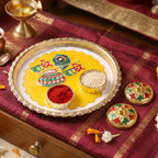 A silver and yellow Radhe Radhe pooja thali with kumkum and rice, next to two decorative containers on a maroon cloth.