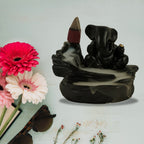 A black Ganesha backflow incense burner with smoke, surrounded by pink flowers and sunglasses on a light blue surface.
