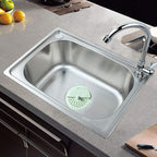 A light green silicone drain strainer sits in a clean stainless steel kitchen sink with a faucet.