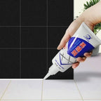 A hand applies white sealant from a tube to the grout line between white floor tiles and black wall tiles.