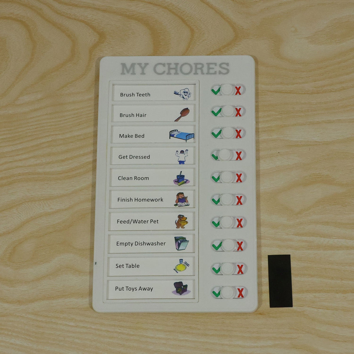 A white chore board with all tasks marked complete, resting on a light wooden surface next to a black rectangular magnet.