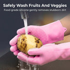 Hands in pink silicone scrubber gloves washing a potato under running water in a kitchen sink.