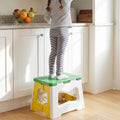 A child stands on a green, white, and yellow plastic stool to reach items on a kitchen counter.