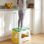 A child stands on a green, white, and yellow plastic stool to reach items on a kitchen counter.
