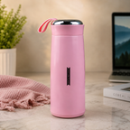 A pink vacuum flask with a silver lid and strap on a marble desk with a laptop and plant in the background.