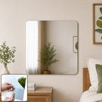 A square mirror with rounded corners hangs above a wooden nightstand in a bedroom, with a small plant