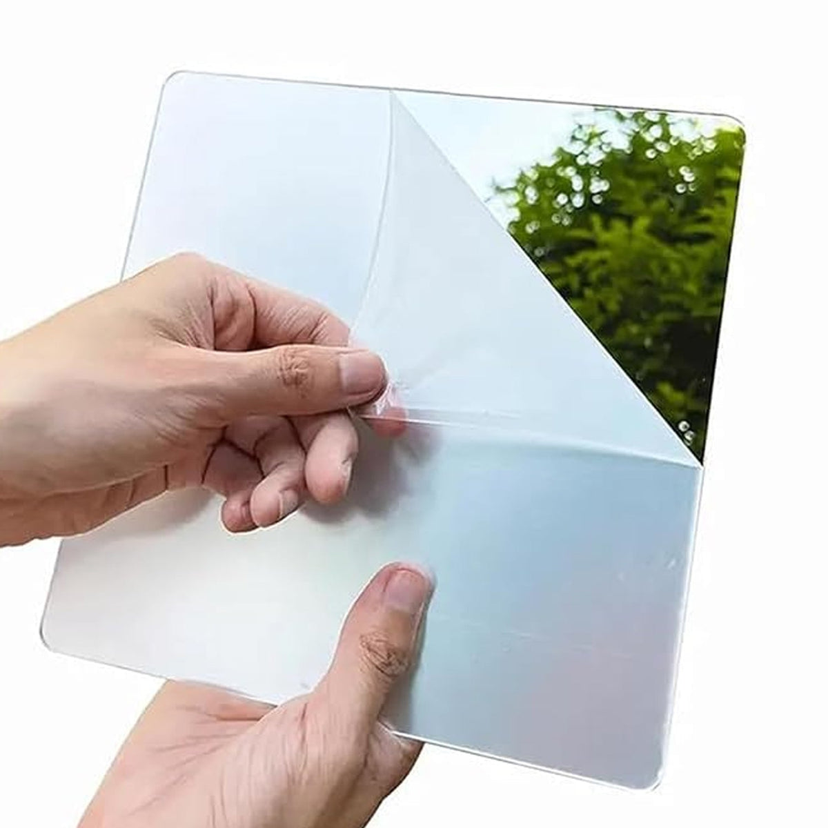 Hands peel back a clear protective film from the surface of a square mirror, reflecting green trees.