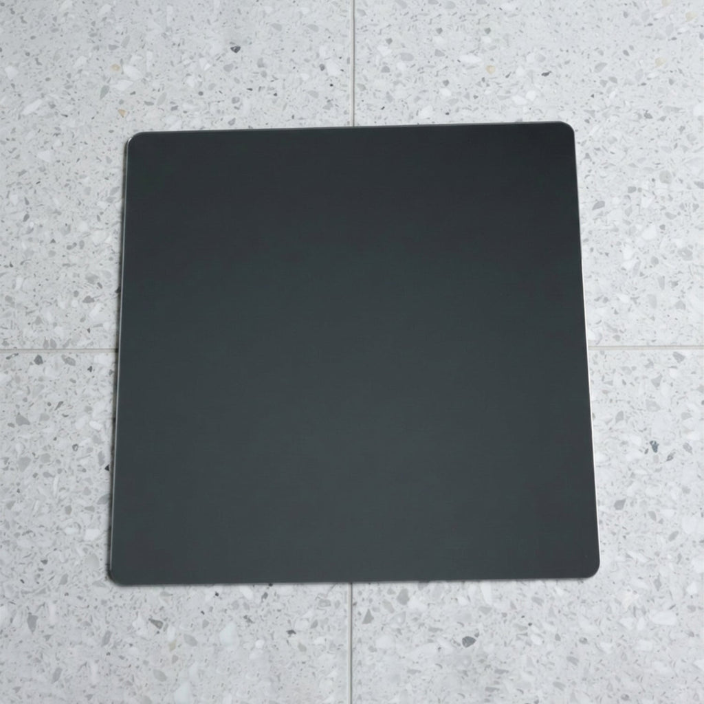 A square mirror with rounded corners lies flat on a speckled gray and white tiled floor.
