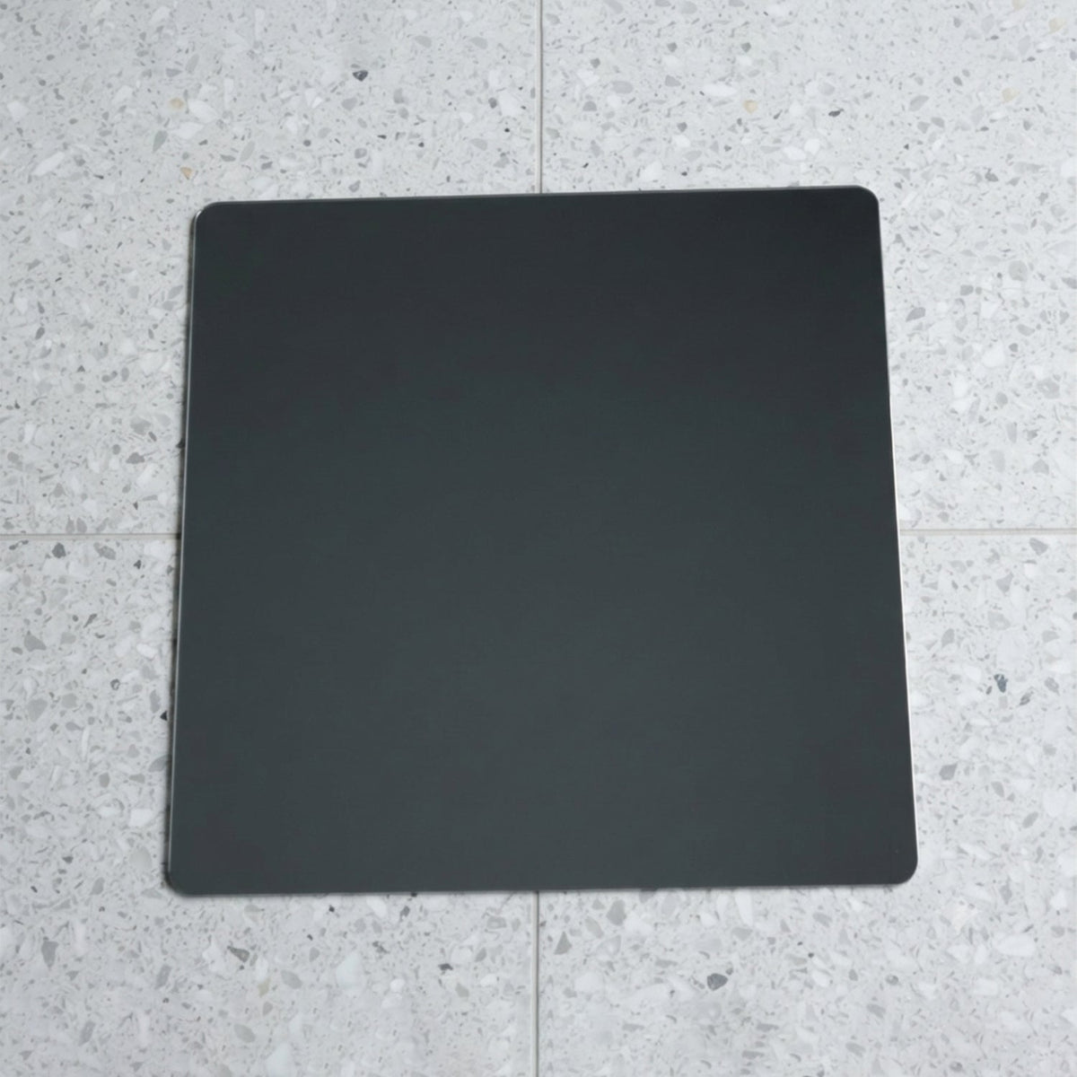 A square mirror with rounded corners lies flat on a speckled gray and white tiled floor.