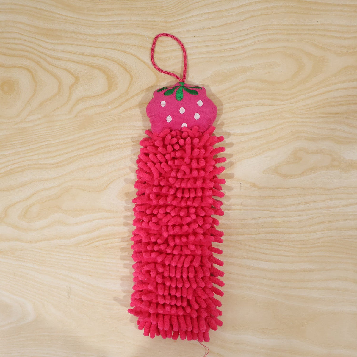 A pink microfiber hand towel with a strawberry top and hanging loop is displayed on a light wooden background.