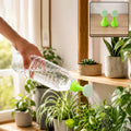 A hand holds a clear plastic bottle with a green and white sprinkler nozzle, watering indoor plants.