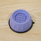 A purple anti-vibration pad with a black rubber base and a grid pattern on its surface, resting on a light wooden background.