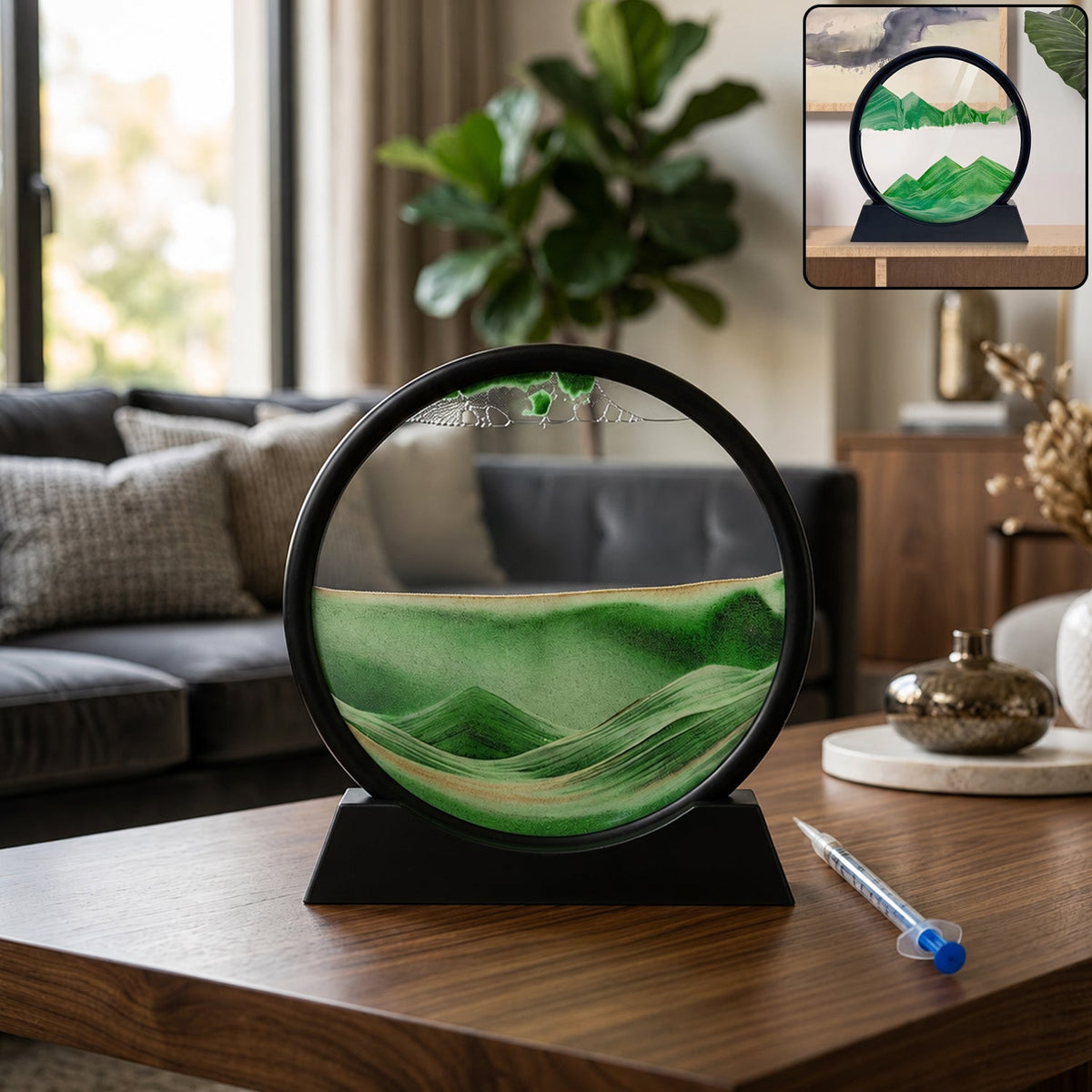 A black-framed circular sand art display with green and gold sand creating a landscape, sitting on a wooden table.