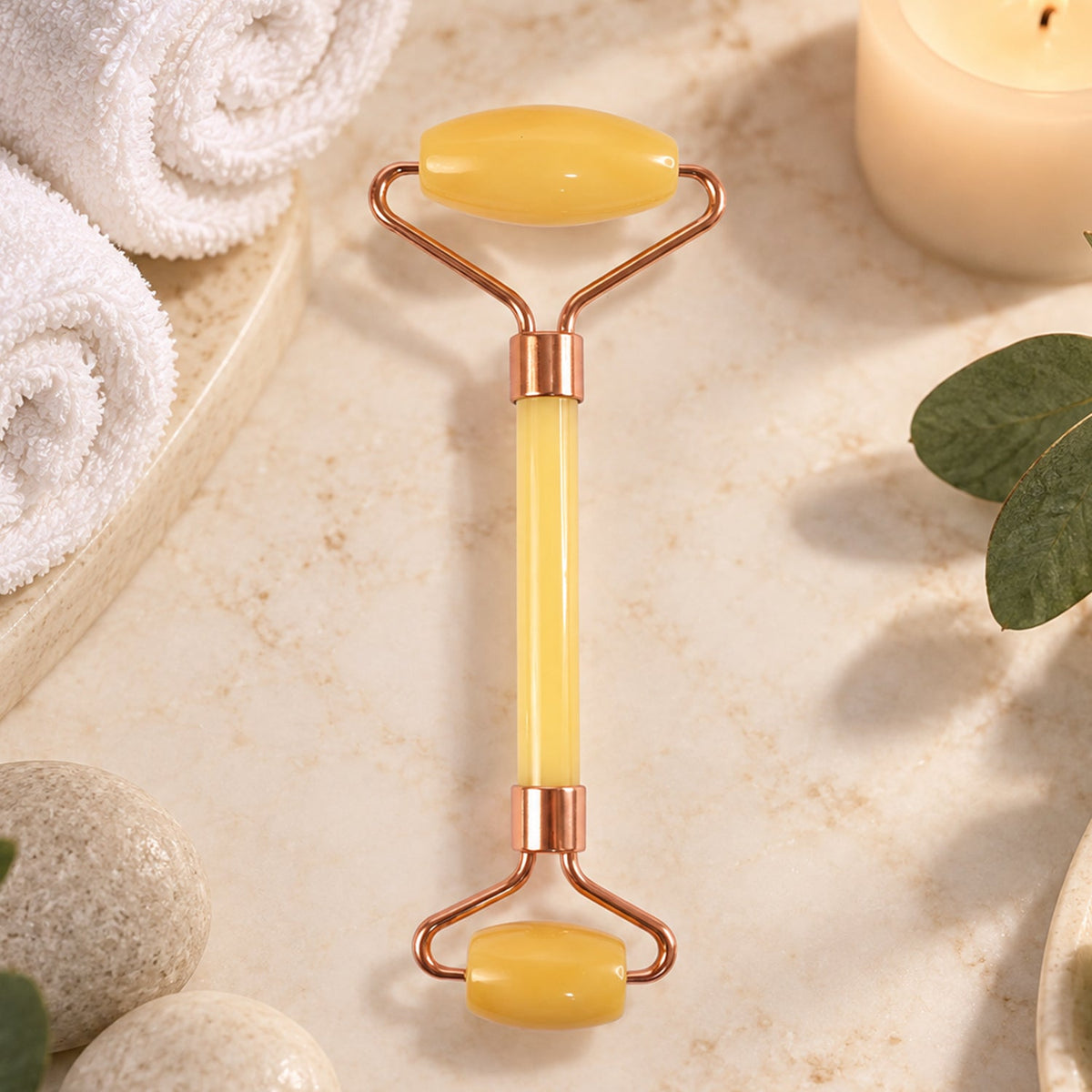A yellow jade facial roller rests on a light-colored marble surface, surrounded by white towels, smooth stones, and a lit