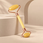 A yellow jade facial roller with rose-gold frame is angled on a light beige background with geometric blocks.