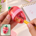 A person's hands sharpen a yellow pencil in a pink and red strawberry-shaped sharpener, creating wood shavings.