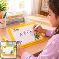 A young girl with dark hair writes “ABC” on a yellow-framed Pexon writing board with a white marker. A small yellow eraser