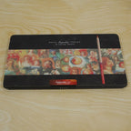 A closed black metal tin with a red colored pencil resting on top, displayed on a light wooden surface.