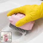 A yellow-gloved hand holds a pink bear-shaped soap dispensing brush, scrubbing a white sink with suds.