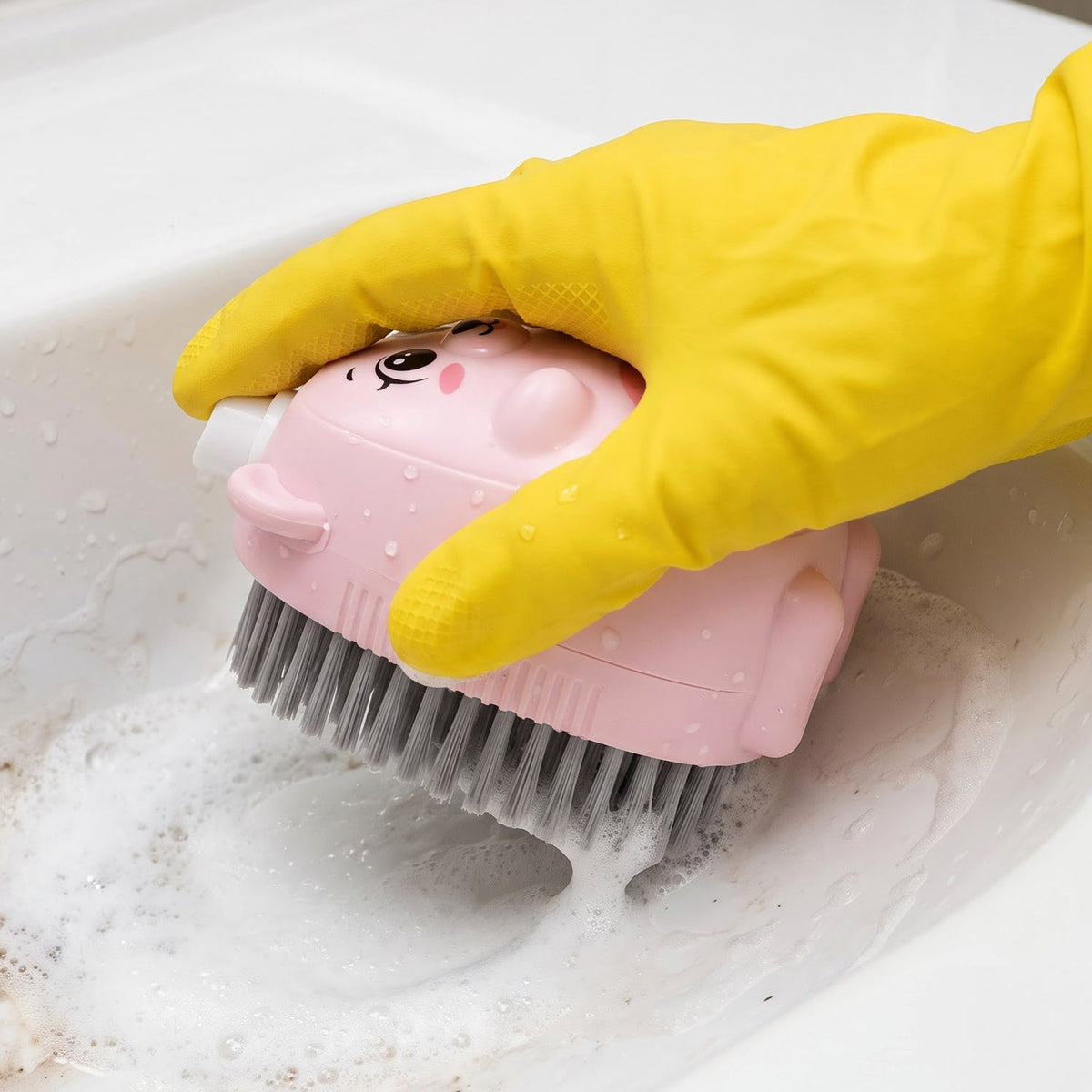 A yellow-gloved hand scrubs a white sink with a pink bear-shaped soap dispensing brush, creating suds.