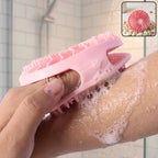 A hand holds a pink silicone body scrubber, lathering soap on an arm in a shower with a small inset image of the scrubber.