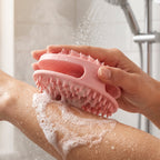 A hand holds a pink silicone body scrubber, scrubbing a soapy arm under a shower head.