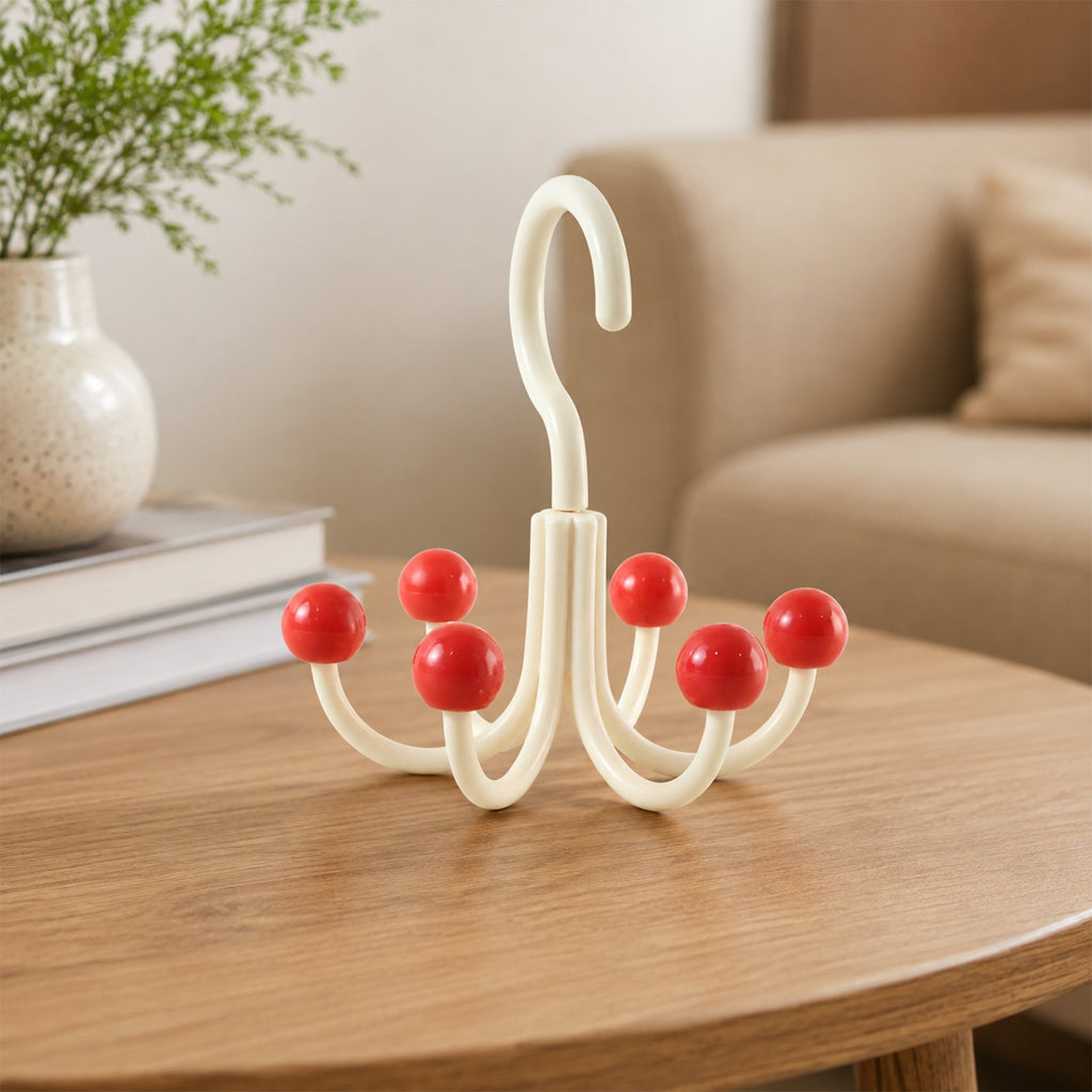 A cream multi-hook hanger with red ball tips sits on a wooden table next to a white vase with greenery, with a beige couch