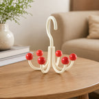 A cream multi-hook hanger with red ball tips sits on a wooden table next to a white vase with greenery, with a beige couch