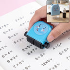 A hand holds a blue elephant-shaped math roller stamp, printing multiplication problems on a white notebook. A small inset
