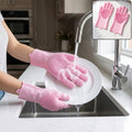 Hands wearing pink silicone gloves with scrubbing bristles washing a white plate under running water in a kitchen sink.