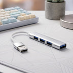 Silver 4-port USB hub with a white cable on a white desk with a keyboard and plant in the background.